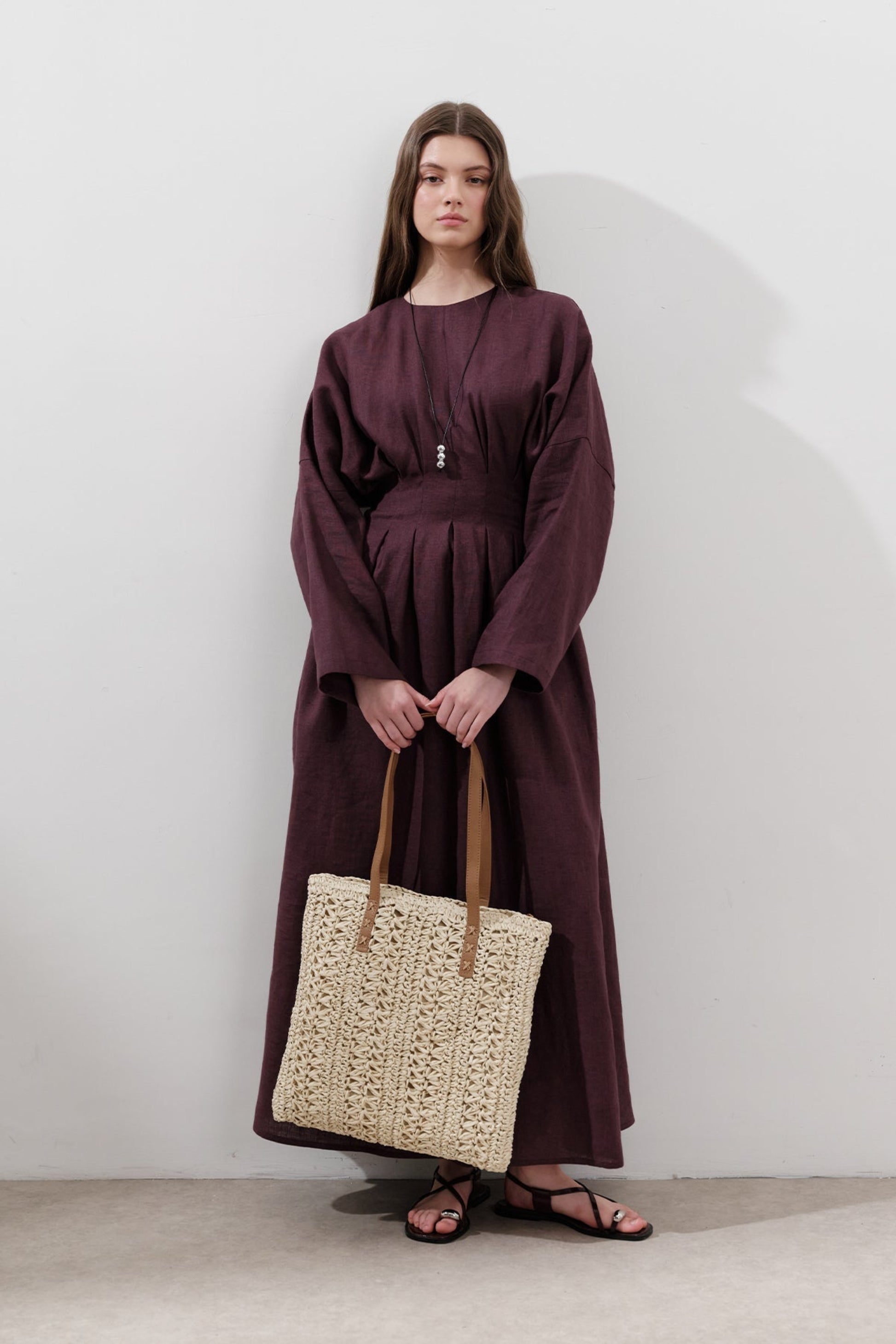 Woman in a long burgundy dress holding a woven beige bag against a white background