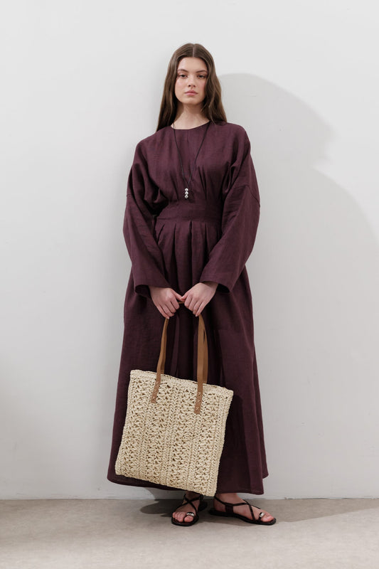 Woman in a long burgundy dress holding a woven beige bag against a white background